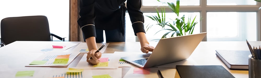 A woman leans over a desk with her laptop open to work on her business plan.