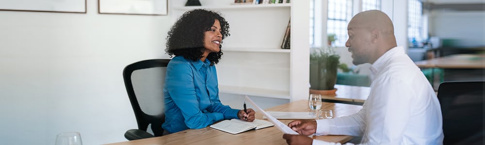 A prospective employee holds his resume and is discussing it as he sits at a desk with the manager.